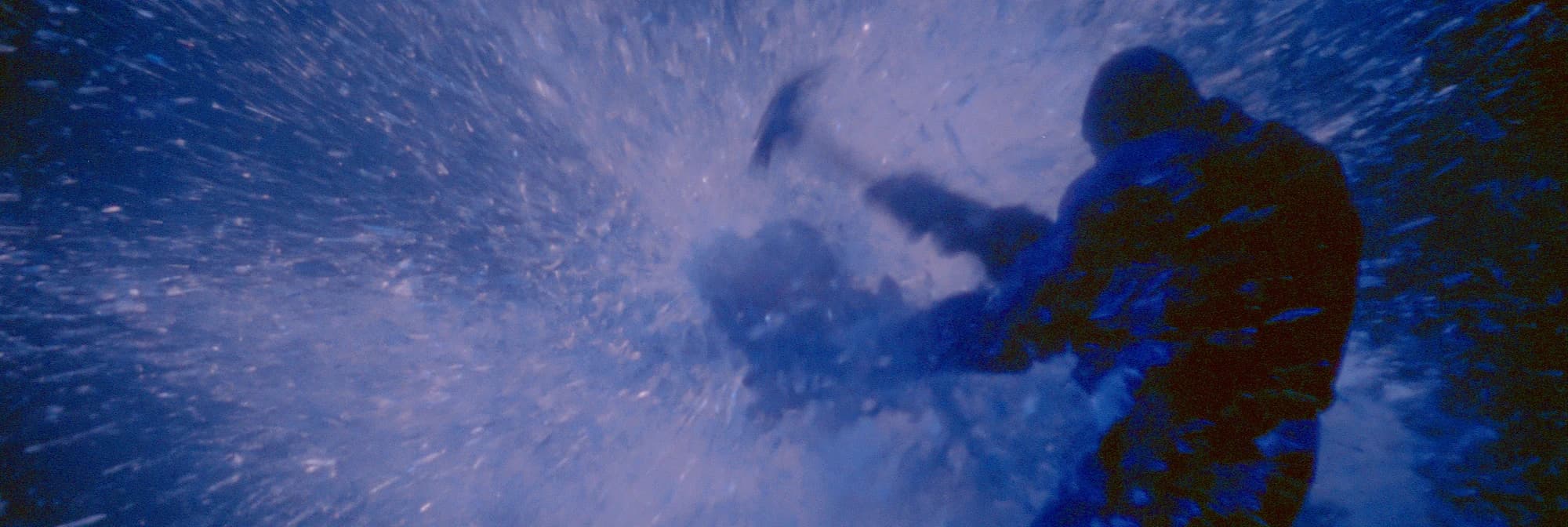 A figure in dark clothing swings a hammer into a burst of shards and spray, all in blue light.