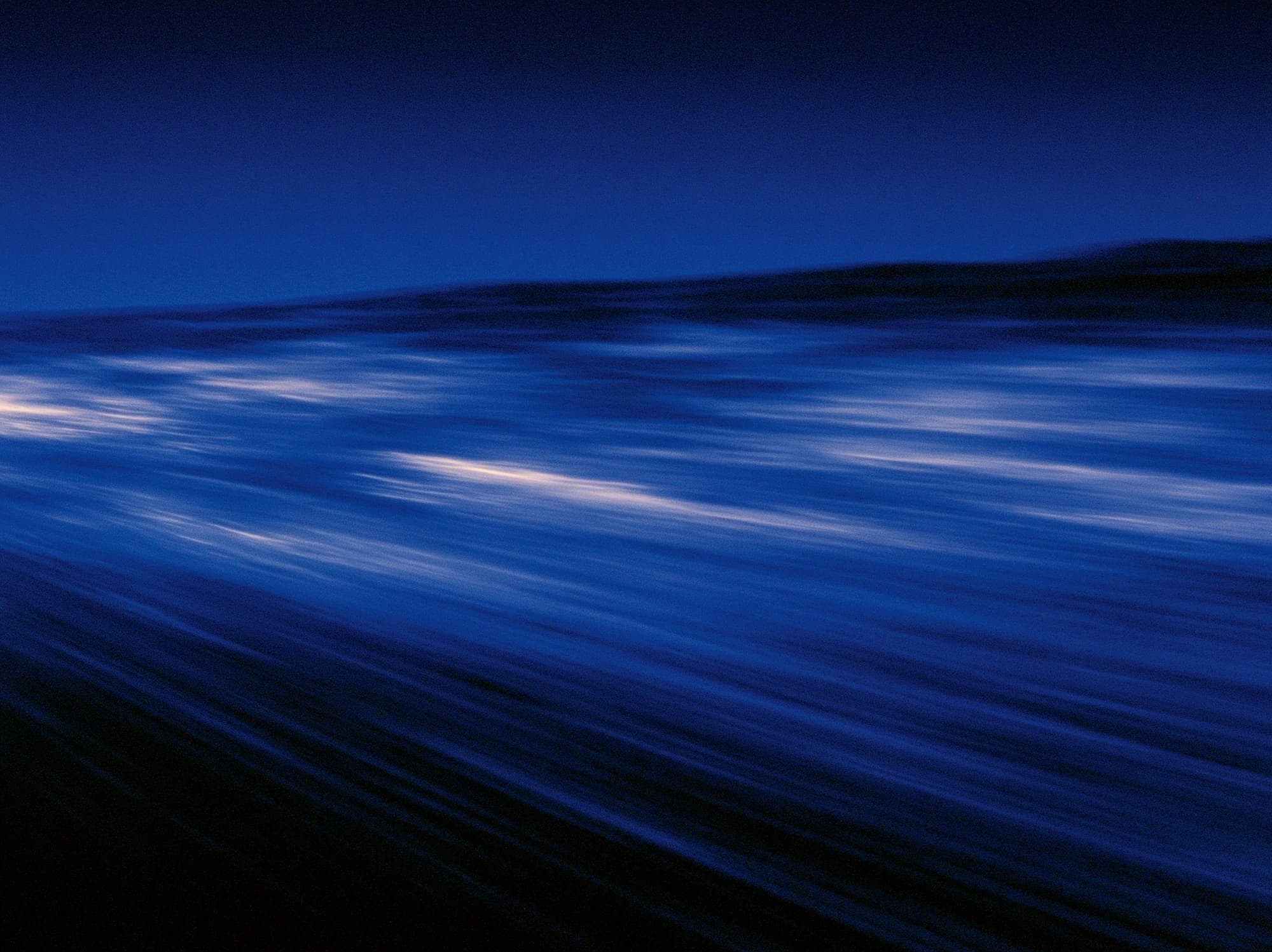 Motion-blurred dark blue water at dusk, with streaks of pale light across the waves and a low ridge on the horizon.