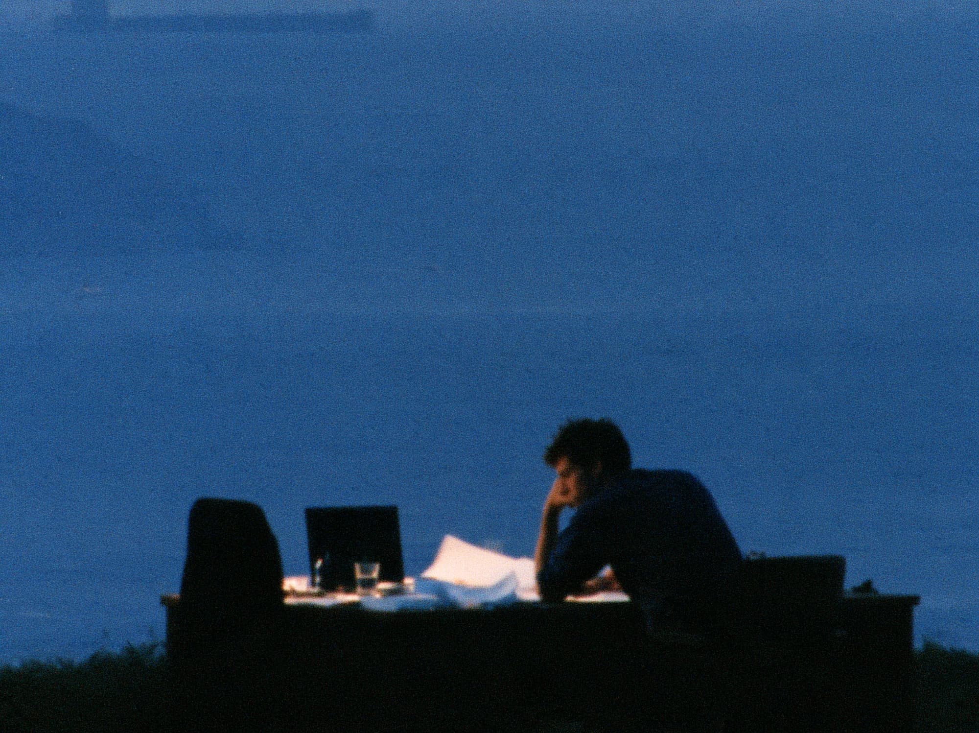 A person sits at a table with papers, resting their head on one hand, in a room filled with blue light.