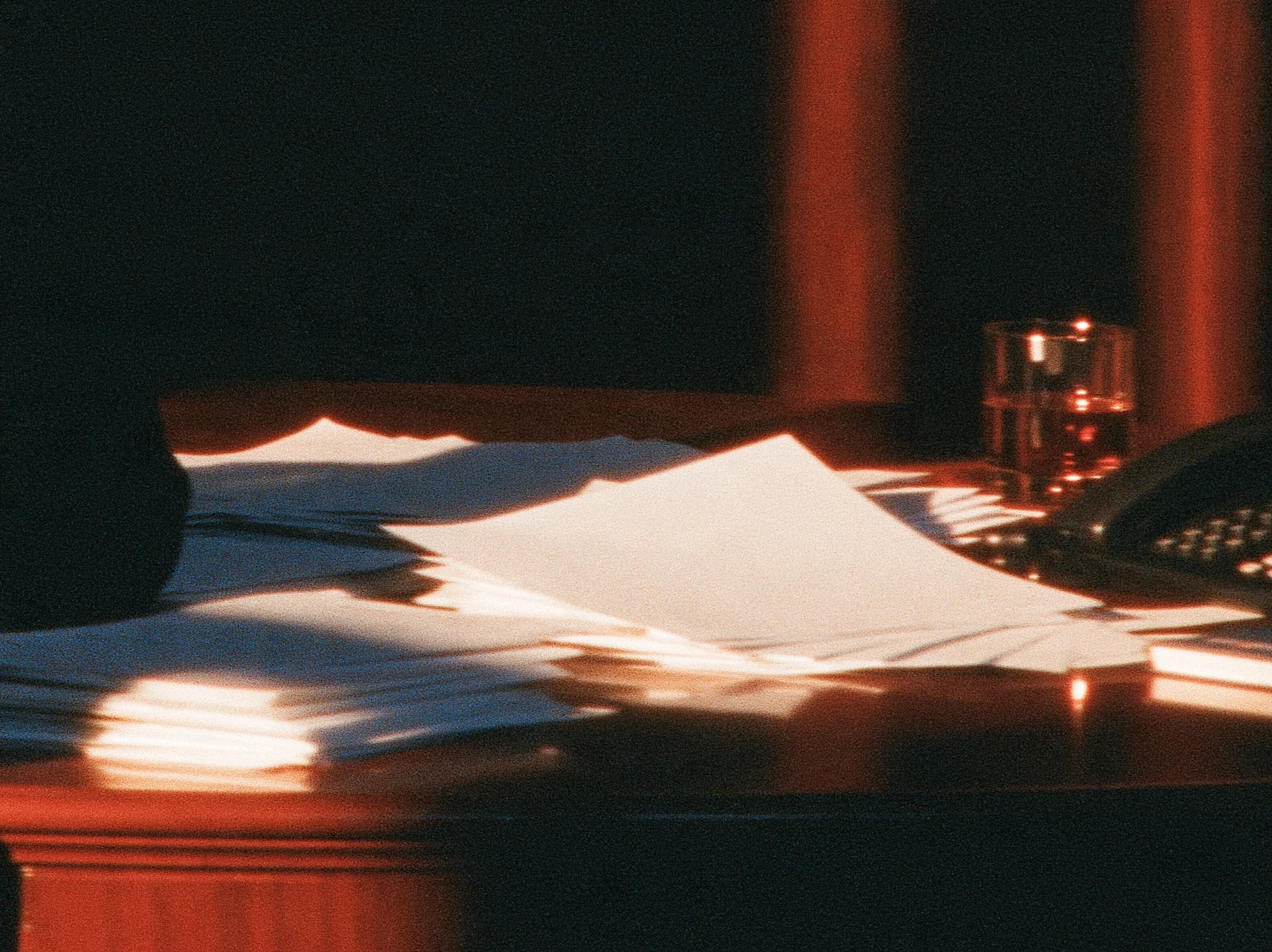 Papers lie across a dark desk in warm orange light, beside a glass and telephone.